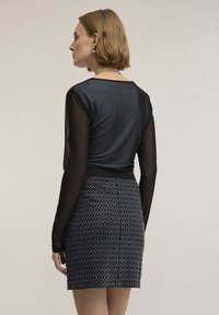 Žena s krátkými světle hnědými vlasy, oblečená v černém transparentním topu s dlouhým rukávem a černé sukni s bílým diamantovým vzorem, stojící nalevo.