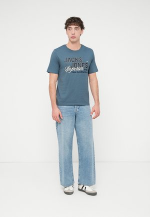 Jeune homme portant un t-shirt graphique bleu, un jean large bleu clair et des baskets blanches, debout devant un fond blanc uni.