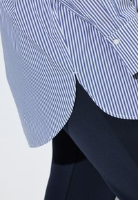 Koszula na guziki w niebiesko-białe paski z zaokrąglonym dołem, noszona na ciemnych legginsach. Wykonana z lekkiego materiału o przemyślanej konstrukcji.