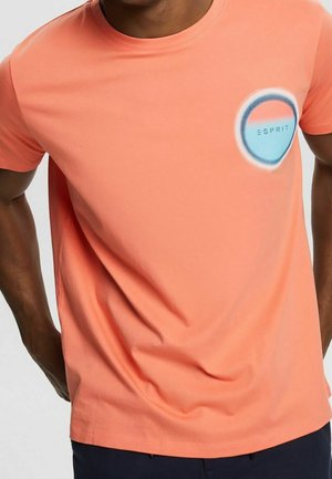 Man draagt een koraalkleurig T-shirt met korte mouwen en een blauw-wit rond Esprit-logo op de bovenste linkerborst.