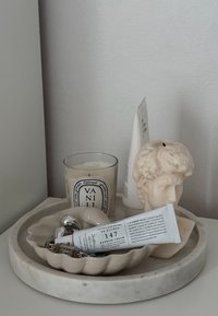 Marmorbrett med bølgende kanter som holder et lys, kremtube, metalobjekter og en figur av et hode. Nøytral fargepalett, glatte teksturer.