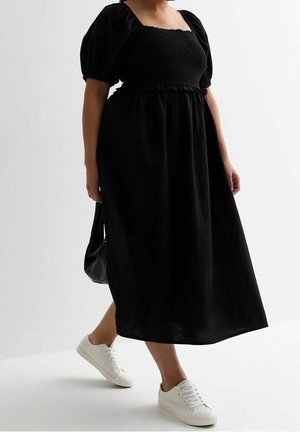 Woman wearing a black puff-sleeve midi dress with smocked bodice, white sneakers, and holding a black handbag walking against a white background.