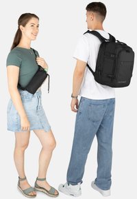 Schwarzer Rucksack mit mehreren Taschen; glatte Textur, rechteckige Form. Model trägt ein eng anliegendes dunkelgrünes Oberteil und einen Jeansrock, hält eine ähnliche Tasche.