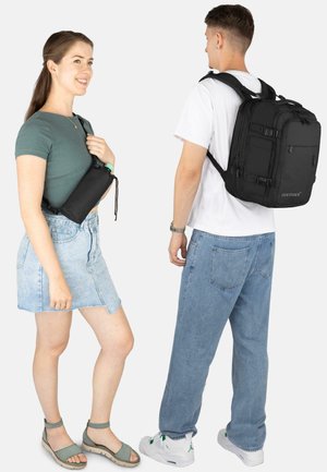 Black backpack with multiple pockets; smooth texture, rectangular shape. Model wearing a fitted dark green top and denim skirt, holding a similar bag.