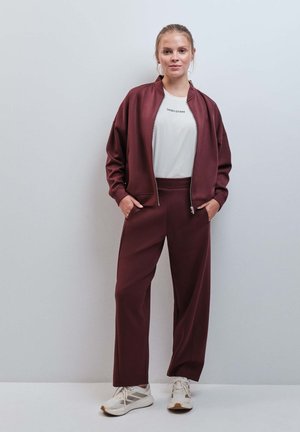 Femme debout les mains dans les poches, portant un survêtement bordeaux, un T-shirt blanc avec un texte et des baskets blanches sur un fond uni.
