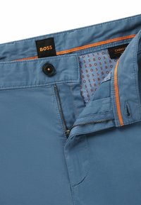Light blue chinos with a smooth texture, featuring a zip fly, button closure, and orange accents on the waistband and pocket zippers.