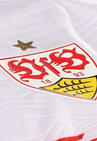 Maglia sportiva bianca con un emblema rosso e oro. L'emblema presenta un design stilizzato con una stella sopra e dettagli neri.