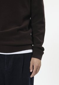 Maglione marrone con texture e scollatura tonda, con polsini a costine, indossato sopra una camicia bianca. Abbinato a pantaloni di velluto a coste blu navy.