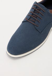 Blue canvas shoe with a rounded toe, brown laces, and a white rubber sole. Textured fabric and contrasting brown accents at the collar.