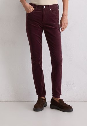 Person iført tætsiddende mørkerøde manchesterbukser og mørkebrune slip-on sko mod en ensartet lys væg.