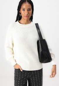 Cremefarbener Strickpullover mit gerippten Bündchen und Saum, kombiniert mit einer schwarzen Leder-Umhängetasche. Das Model trägt dunkle Jeans mit silbernen Nieten.