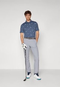 Golfový outfit pozostávajúci z tmavomodrej polokošele s vzorom, svetlosivých nohavíc, sivého opasku, bielych golfových topánok s modrými akcentmi a golfovej palice v ruke.