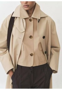 Béžový trench coat přes béžovou košili na knoflíky s hnědými knoflíky a límcem. Tmavě hnědé kalhoty s detaily na stažení.