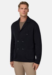 Tumes sinine topelt-kaluga jakk, mis on valmistatud tekstureeritud kangast, suured mustad nööbid ja kaks esist pihustus. Kantud helepruuni särk üle.