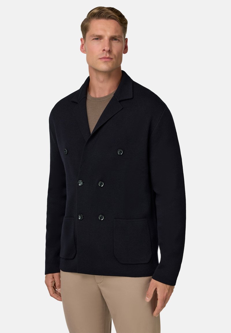 Tumes sinine topelt-kaluga jakk, mis on valmistatud tekstureeritud kangast, suured mustad nööbid ja kaks esist pihustus. Kantud helepruuni särk üle.