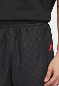 Μαύρο αθλητικό σορτς με υφή ζιγκ-ζαγκ, ελαστική μέση και κόκκινο διακριτικό λογότυπο στο πλάι. Το υλικό φαίνεται ελαφρύ.