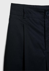 Pantalon noir à devant plat, doté d'un seul pli, d'une fermeture par bouton et d'une ceinture robuste. Le tissu est lisse avec un léger éclat.