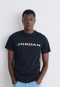 Melnas kokvilnas T-krekls ar "JORDAN" logo baltā krāsā un sarkanām akcentēm. Īsas piedurknes, apaļa kakla izgriezuma dizains, valkāts ar gaiši ziliem džinsiem.