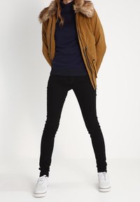 Brun vadderad jacka med en fuskpälsfodrad huva, lager på lager över en marinblå sweatshirt, tillsammans med svarta skinny jeans och vita sneakers.