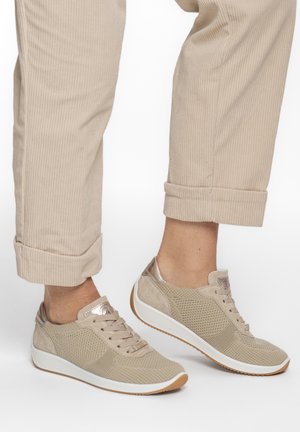 Beige gebreide sneakers met witte zolen gedragen bij beige corduroy broeken die bij de enkels zijn opgerold, op een witte achtergrond.