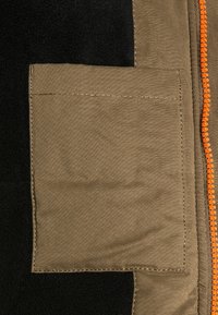 Kieszeń w kolorze khaki z przeszytymi krawędziami na czarnym tle materiału, z pomarańczowym suwakiem po prawej stronie.