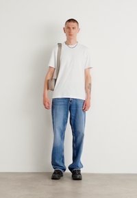 T-shirt blanc à manches courtes, jeans en denim bleu, chaussures noires, sac à épaule gris, posé contre un fond blanc uni.