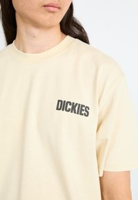 Beige bomulds t-shirt med rund hals, korte ærmer og et sort trykt logo, der står "DICKIES" på venstre side.