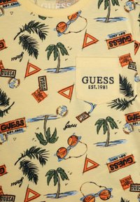 Camiseta de algodón amarilla con un estampado tropical que presenta palmeras, gafas de sol y logotipos de marca. Incluye un bolsillo frontal con bordado.