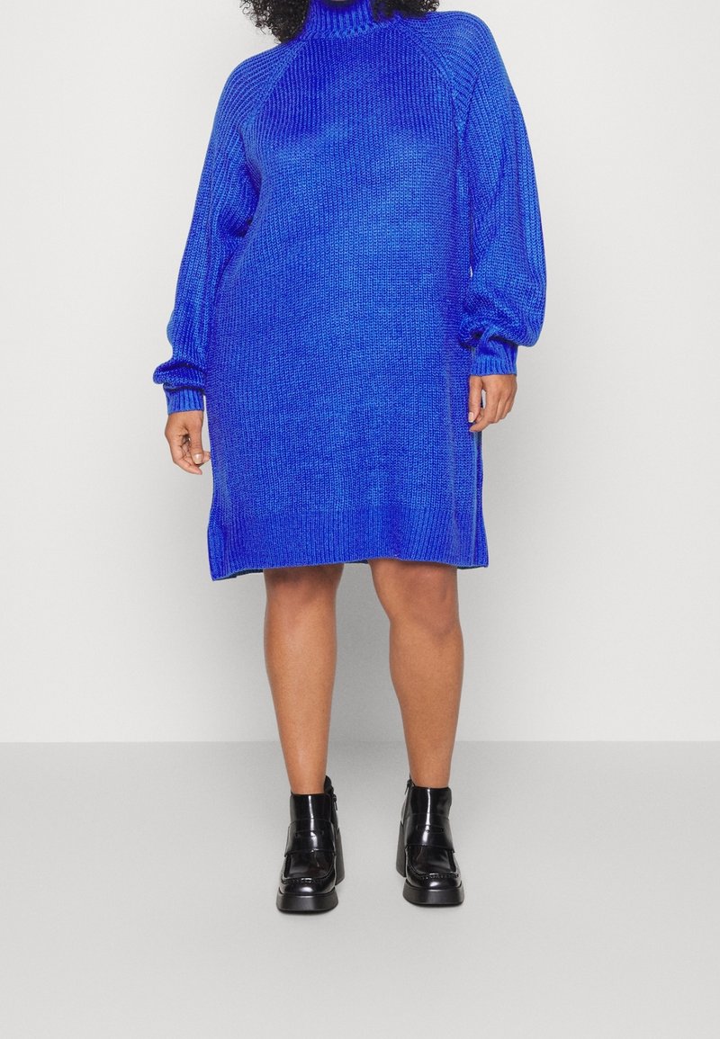 Femme portant une longue robe en tricot bleu roi avec des manches ballon et des bottines noires, debout devant un fond uni.