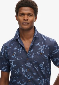 Camisa de botón de manga corta en azul marino oscuro con un patrón floral en azul claro, dos bolsillos en el pecho y botones de madera. Tejido texturizado.