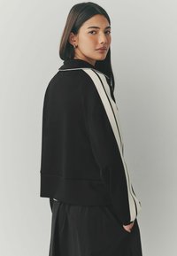 Sweatshirt noir avec un col montant, présentant des accents rayés blancs le long des bras. Texture douce, design court et coupe décontractée.