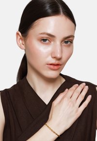 Bracelet en or torsadé porté au poignet, contrasté avec un vêtement texturé brun. La peau du modèle apparaît lisse avec une luminosité naturelle.