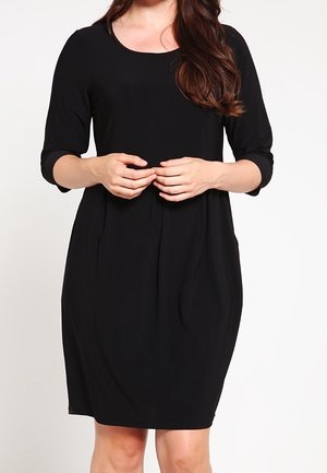 Femme portant une robe noire simple jusqu'aux genoux, avec des manches trois-quarts, debout les mains doucement jointes devant elle.