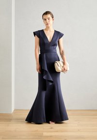 Robe de soirée bleu marine avec un décolleté en V profond, des manches courtes, une taille cintrée et un ourlet évasé. Accompagnée d'un sac à main crème avec des anses en chaîne.