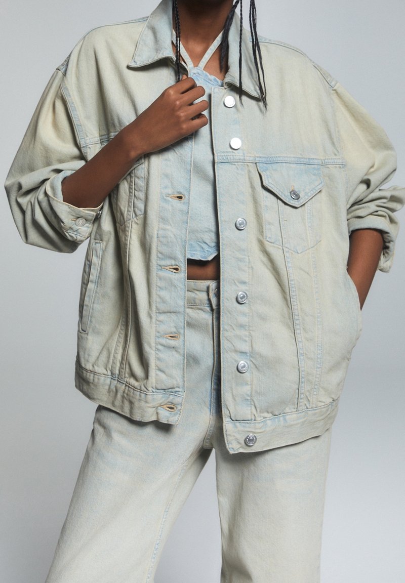 Chaqueta de mezclilla azul claro con un corte holgado, botones, bolsillos delanteros y un ligero desvanecido. Combinada con pantalones de mezclilla a juego.