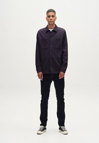 Tamsiai violetinė mygtukinė striukė su dviem priekinėmis kišenėmis, dėvima ant baltos marškinėlių, derinama su tamsiomis džinsinėmis kelnėmis ir juodais sportbačiais.