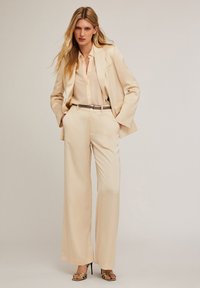Krämfärgad kostym med en strukturerad kavaj och vida byxor. Innehåller en ljusbeige skjorta och ett metalliskt bälte. Slät tyg med en skräddarsydd passform.