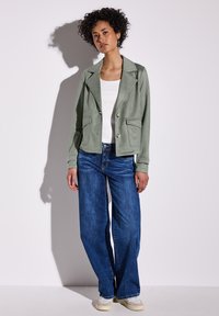 Grüner cropped Blazer mit Revers, zwei vorderen Taschen und goldenen Knöpfen; kombiniert mit weit geschnittenen blauen Jeans und beigen Turnschuhen.