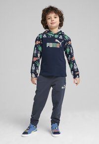 Sudadera navy con estampados coloridos de manos y figuras, logo de PUMA en el centro, combinada con pantalones de chándal gris oscuro y zapatillas azules con detalles del logo.
