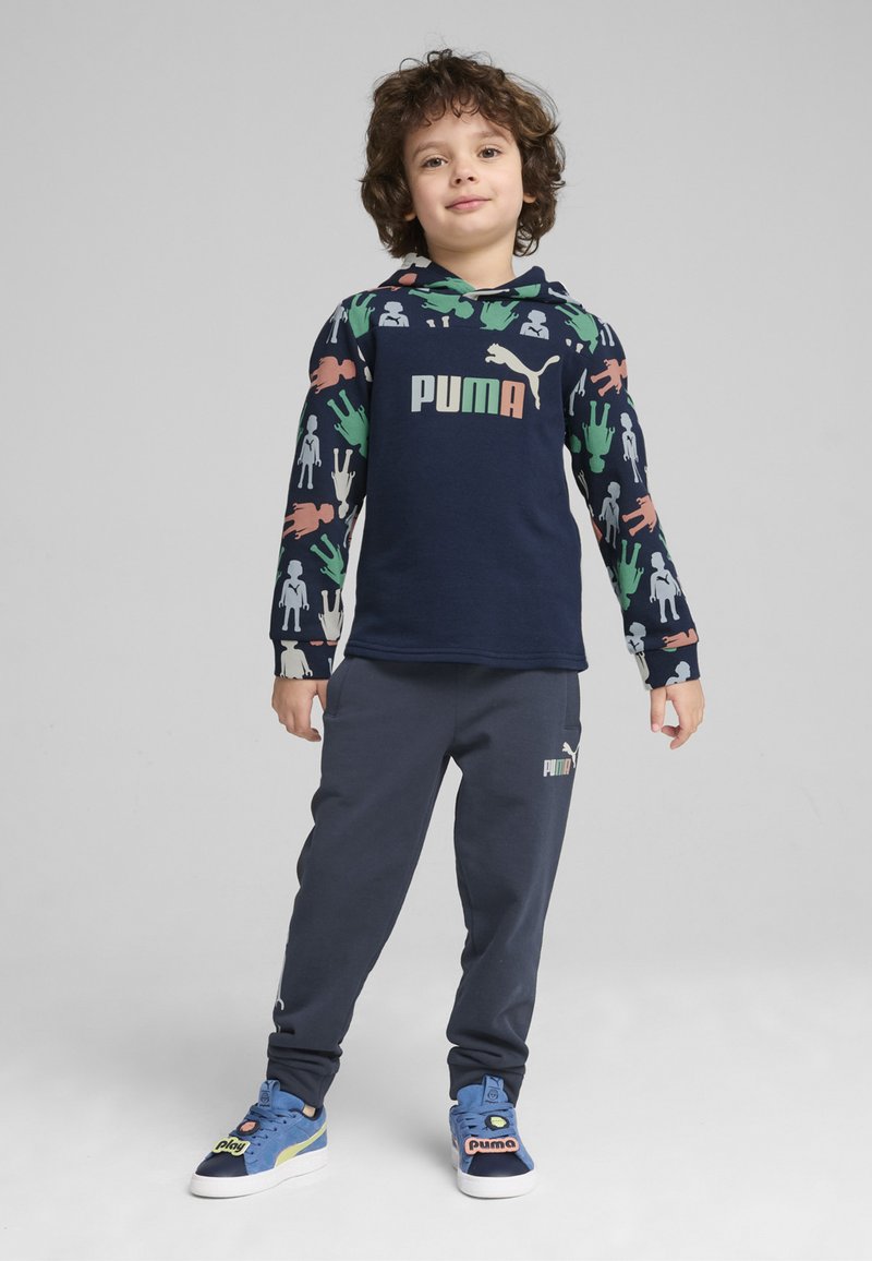Sudadera navy con estampados coloridos de manos y figuras, logo de PUMA en el centro, combinada con pantalones de chándal gris oscuro y zapatillas azules con detalles del logo.