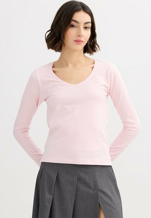 Mujer con cabello corto y oscuro, vistiendo una camiseta de manga larga de color rosa claro con cuello en V y una falda gris plisada sobre un fondo liso.