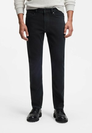 Mann trägt schwarze Slim-Fit-Jeans kombiniert mit schwarzen Leder-Schnürschuhen und einem hellfarbenen Pullover mit langen Ärmeln.