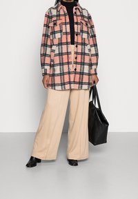 Karo sako v růžové, černé a bílé barvě s dvěma náprsními kapsami, zapínáním na knoflíky a volným střihem, spojené s béžovými kalhotami s širokými nohavicemi a černými botami.