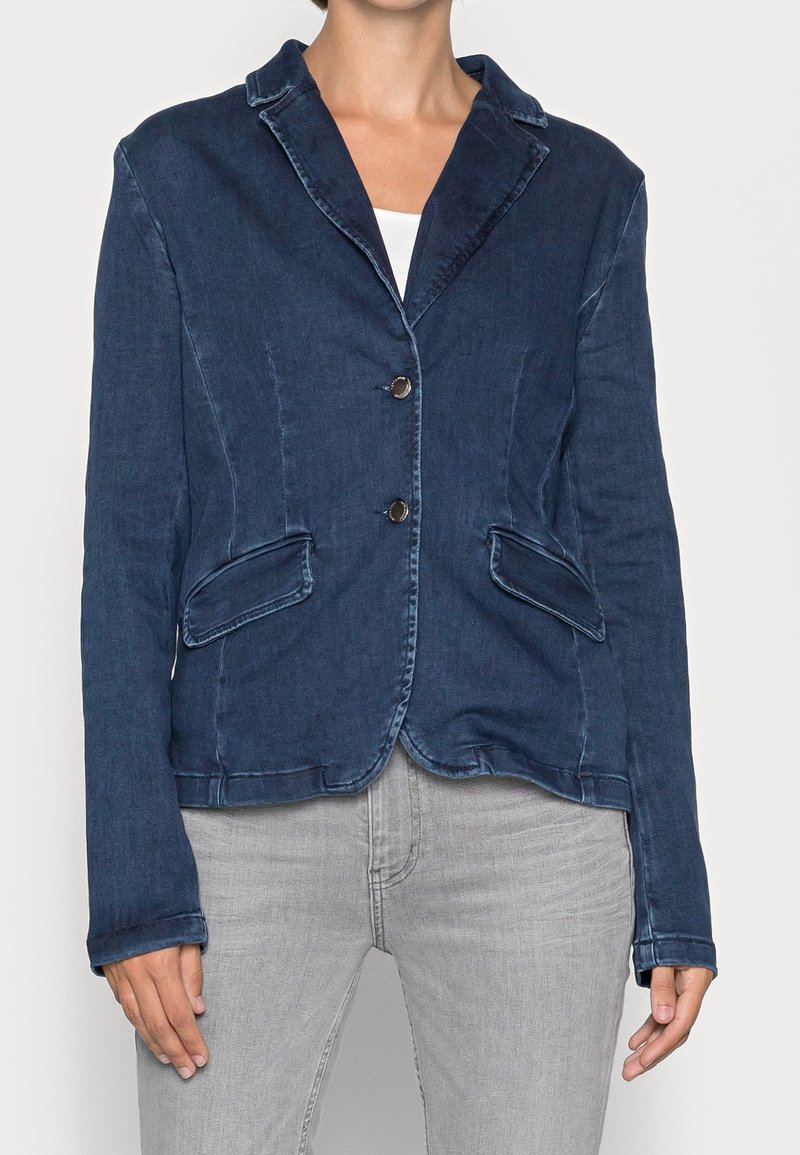 Femme portant un blazer en denim bleu foncé ajusté avec des revers crantés, deux poches avant à rabat et deux boutons, associé à un jean gris clair.