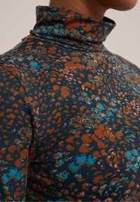 Rolák s abstraktním květinovým vzorem v tmavě modré, rezavě oranžové a tyrkysové barvě na horní části trupu a krku osoby.