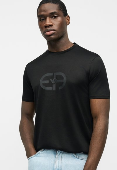 Νέος άνδρας που φοράει μαύρο κοντομάνικο πουκάμισο με διακριτικό ανάγλυφο λογότυπο, ανοιχτό μπλε τζιν, στέκεται με το ένα χέρι στην τσέπη, ουδέτερο φόντο.