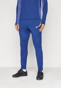 Modré atletické nohavice vyrobené z elastického materiálu s priliehavým strihom, zdobené ružovým logom Nike na boku a čiernymi tréningovými topánkami.