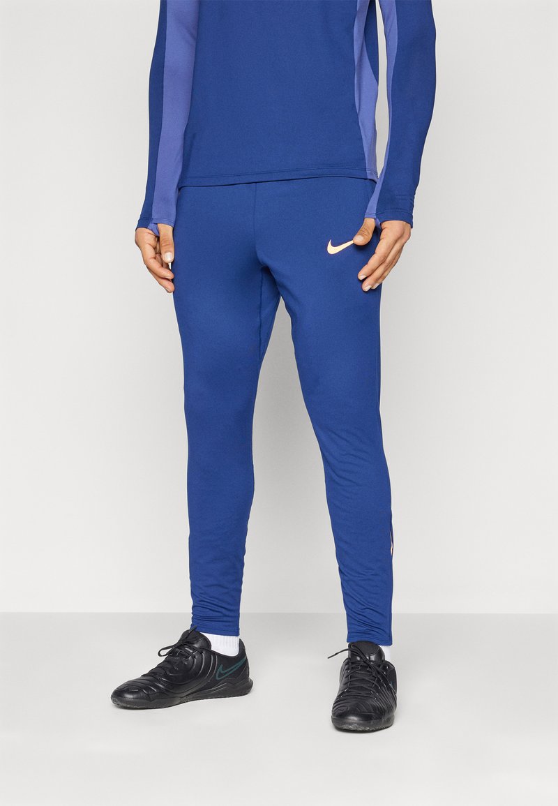 Modré atletické nohavice vyrobené z elastického materiálu s priliehavým strihom, zdobené ružovým logom Nike na boku a čiernymi tréningovými topánkami.