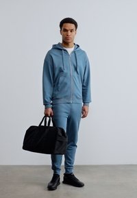 Νεαρός άνδρας με μπλε φόρμα που κρατά μια μαύρη τσάντα duffel, στέκεται μπροστά σε απλό ανοιχτό γκρι τοίχο και τσιμεντένιο πάτωμα.