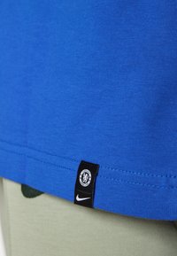 Blå bomullströja med rak skärning, med en svart etikett som har Chelsea FC:s logotyp och en Nike swoosh vid fållen. Bakgrunden är ljusgrön.